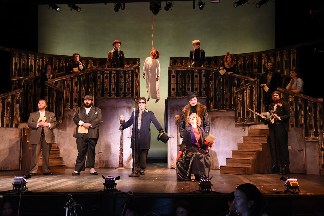 A chorus of people singing onstage, with a woman who was hanged behind them