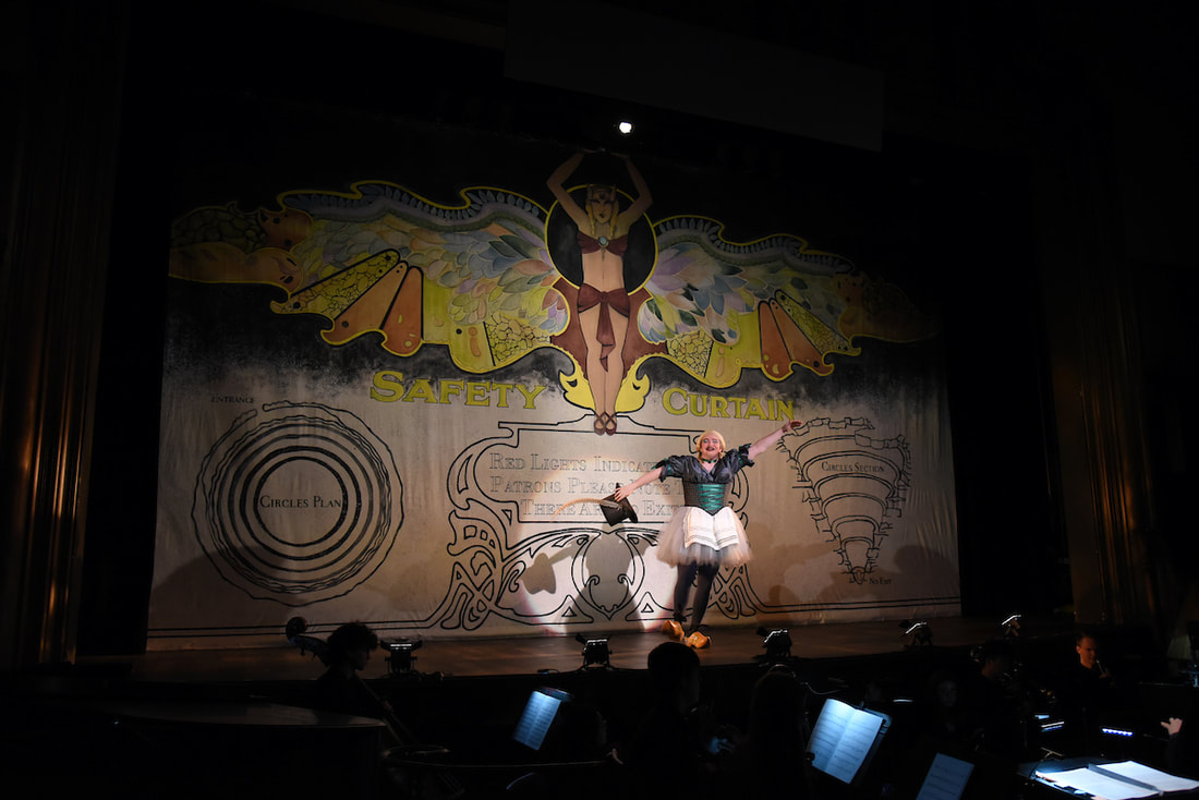 A man dressed as a female maid stands centerstage, singing