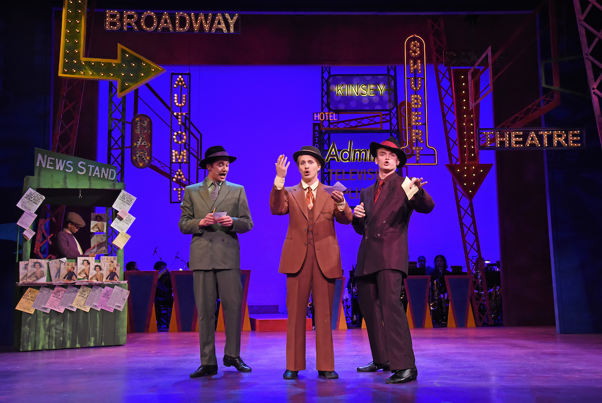 Three men stand onstage singing, with signs of New York behind them