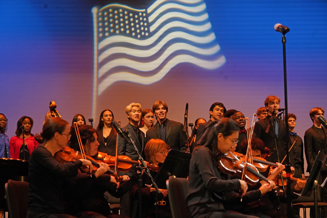 An orchestra and a chorus of singers with a projected image of an American flag in the background