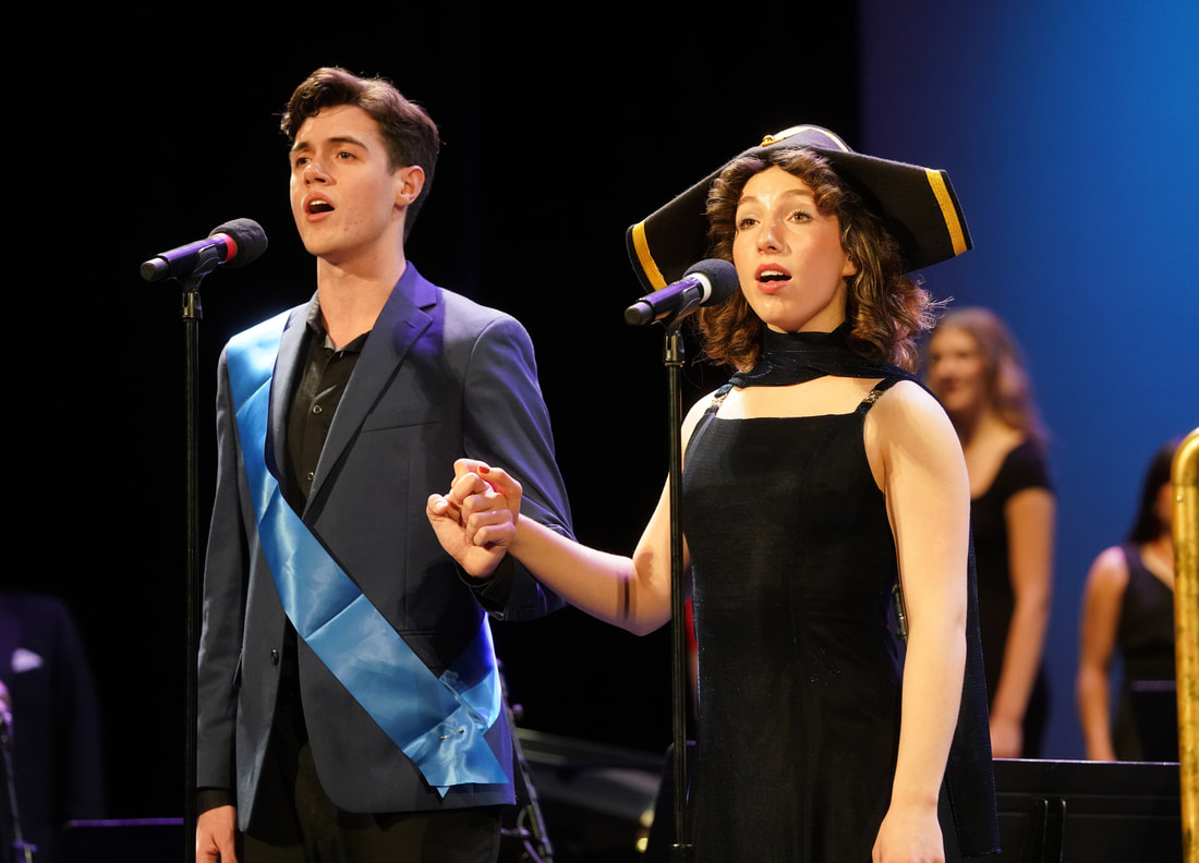 A man and woman stand hand in hand, singing into microphones on stands