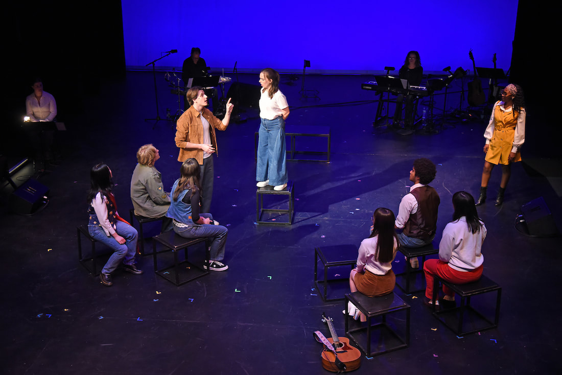 A group of students sit with their backs to the camera facing a girl standing on a block